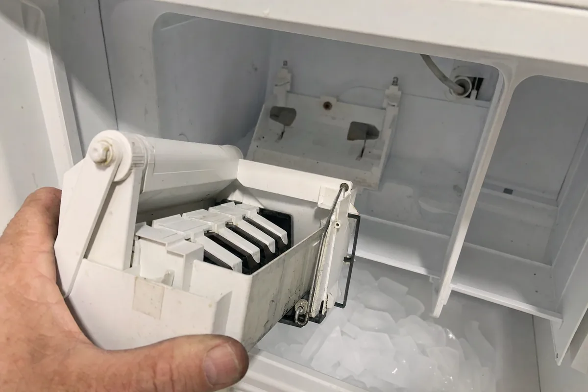 A hand lifting the white ice maker module clear of the freezer compartment, finished cubes lying in the bin below