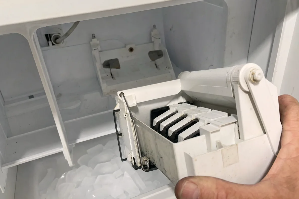 The ice maker module held at an angle in the freezer opening, showing the mould, the ejector arm and the cubes underneath