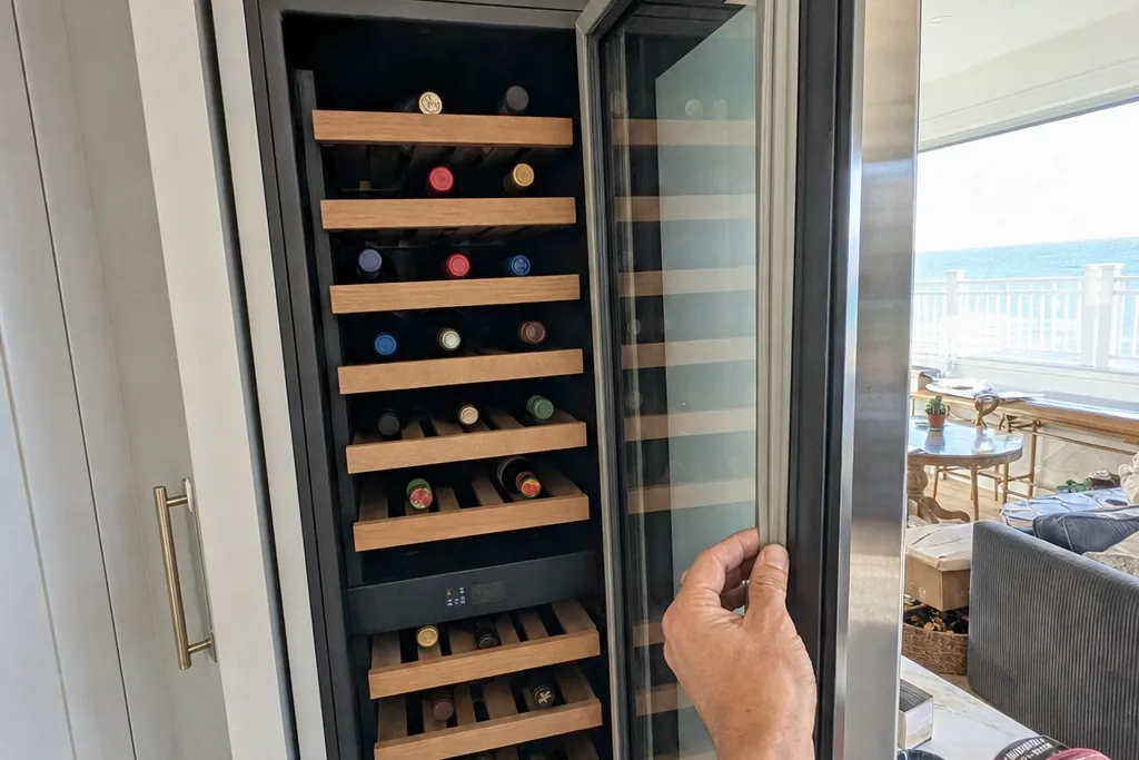 A tall glass-door wine column filled with bottles on wooden racks, standing in a bright kitchen with water visible through the window