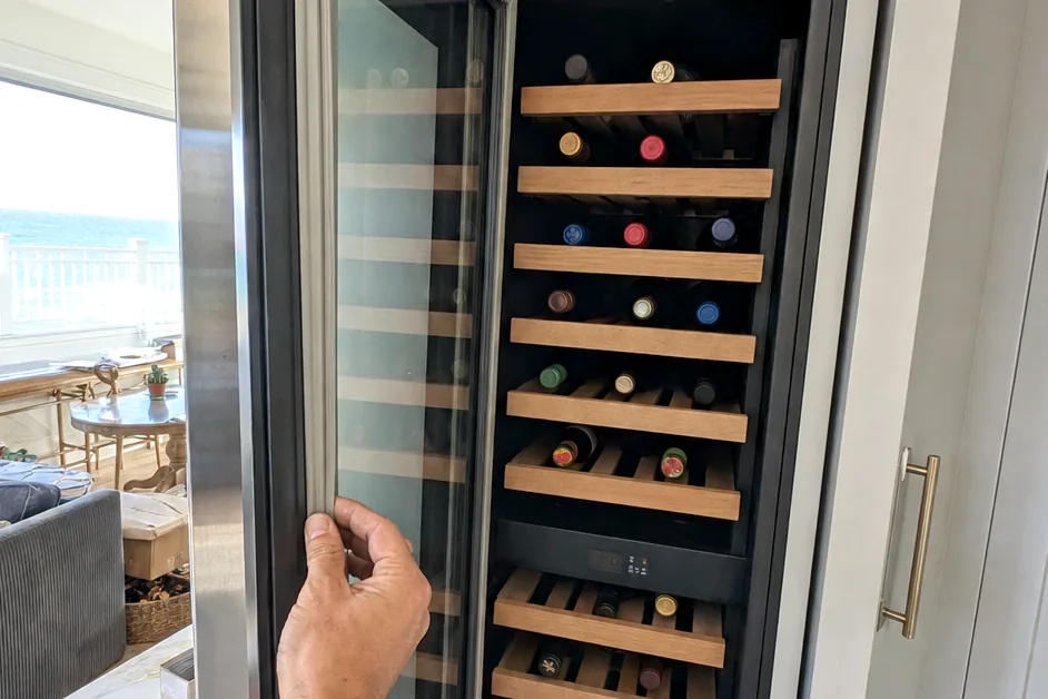 A hand on the handle of a full-height integrated column, its glass door reflecting the cabinetry it is set into