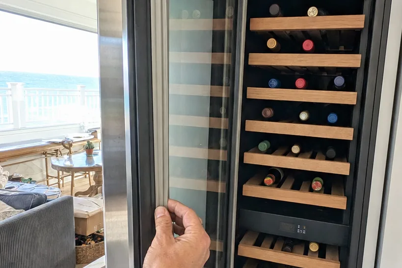A wine column in an island kitchen, daylight off the water falling across its glass door and the bottles inside