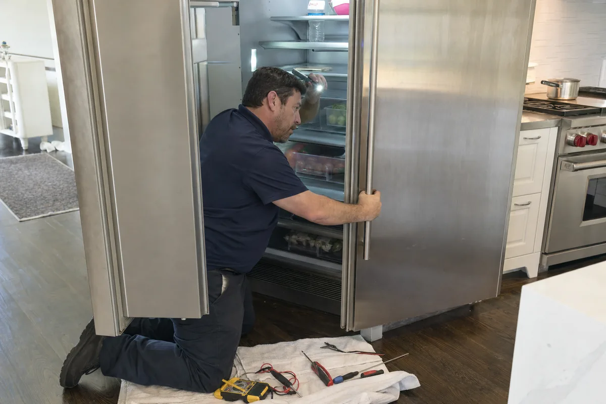 A technician kneeling at an open stainless built-in refrigerator, both doors swung wide, in a kitchen with a range behind him